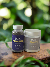 Load image into Gallery viewer, 92+ Parasite Cleanse and Zeolite bottles on a wooden surface with a green blurred background.